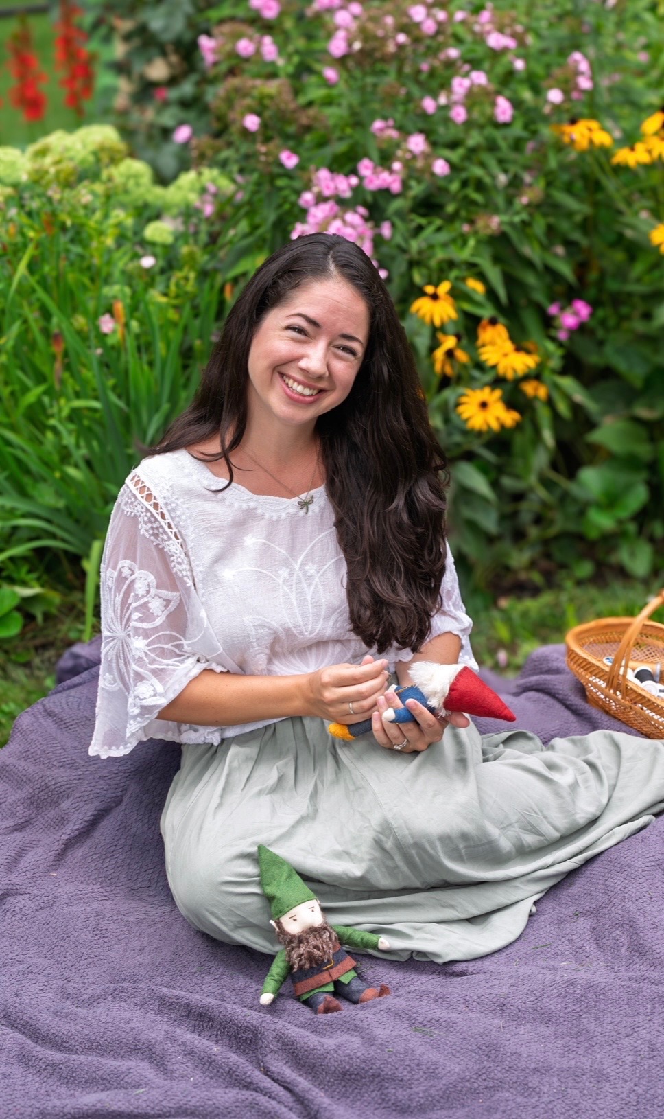 Brooke Kelly smiling in a flower garden while holding handmade dolls.