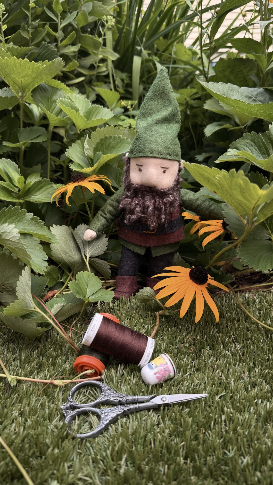 Handmade green-hatted gnome posed among garden flowers with sewing notions.